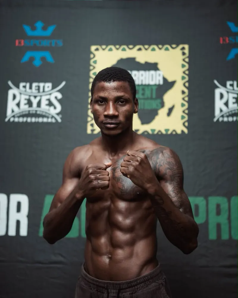 Michael Adesodun faces the camera with both fists raised before a Warrior Ascent backdrop