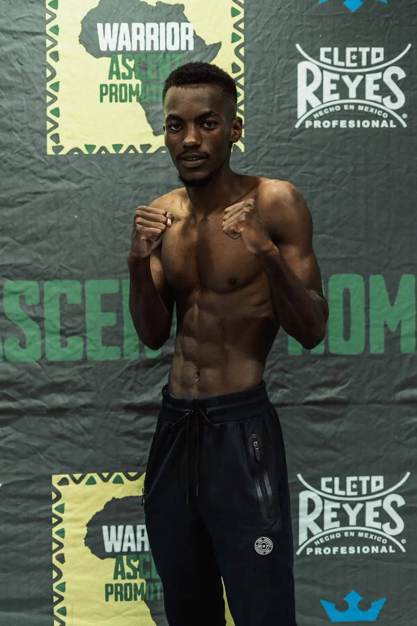 A boxer poses with both fists raised in front of a Warrior Ascent weigh-in backdrop