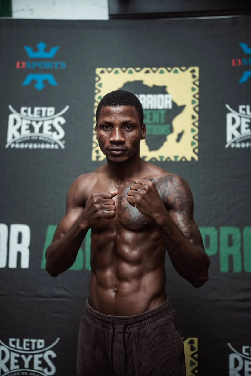 A boxer stands with fists raised in front of a Warrior Ascent weigh-in backdrop