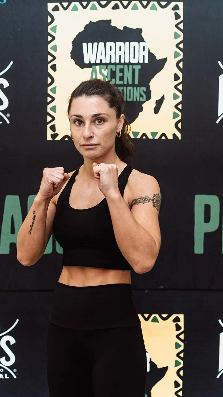 A boxer poses in a guard stance in front of a Warrior Ascent weigh-in backdrop